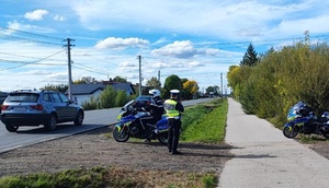 policjanci ruchu drogowego prowadzą pomiar prędkości pojazdów jadących po drodze. Jeden z policjantów siedzi na motocyklu służbowym.