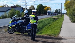 policjanci ruchu drogowego prowadzą pomiar prędkości pojazdów jadących po drodze. Jeden z policjantów siedzi na motocyklu służbowym.