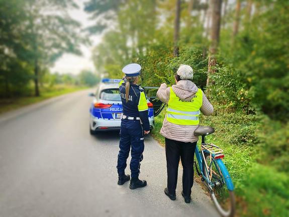 w tle stoi radiowóz, na pierwszym planie policjantka ruchu drogowego przekazuje rowerzystce kamizelkę odblaskową