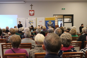 seniorzy na prelekcjach z policjantami. Policjantka w oddali mówi do mikrofonu.