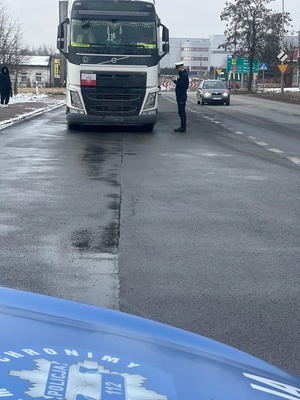 Policjant ruchu drogowego podczas kontroli dokumentów kierowcy pojazdu ciężarowego. Widoczna ciężarówka i maska radiowozu.