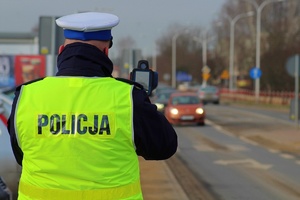 Policjant ruchu drogowego prowadzi kontrole prędkości przy użyciu urządzenia laserowego. W oddali widać nadjeżdżające auta.