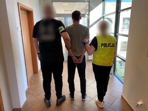 Zatrzymany mężczyzna prowadzony w kajdankach przez policjantów po korytarzu budynku.