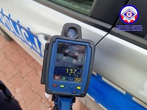 Laserowy miernik prędkości z pomiarem, trzymany w dłoni na tle policyjnego radiowozu.