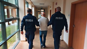 Umundurowani policjanci prowadzą korytarzem zatrzymanego mężczyznę.