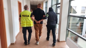 Policjanci prowadzą korytarzem pomiędzy sobą zatrzymanego mężczyznę, który ma nałożone kajdanki na ręce trzymane z tyłu.