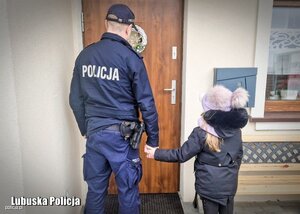 Umundurowany policjant trzyma za rękę dziewczynkę. Obydwoje stoją przed drzwiami.