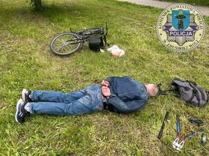 Na trawniku leży twarzą do ziemi mężczyzna, za nim leży przewrócony rower, a koło mężczyzny jest plecak, są rozłożone na trawniku narzędzia: nóż do cięcia metalu, pompka rowerowa, zapięcie od roweru.