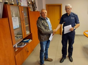 Umundurowany policjant ściska dłoń drugiego mężczyzny i przekazuje mu dokumentację.