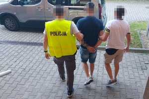 Policjanci prowadzą zatrzymanego mężczyznę z kajdankami na nadgarstkach rąk trzymanych z tyłu do samochodu.