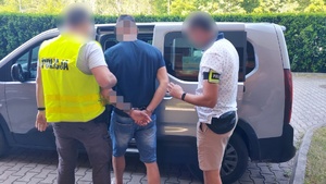 Policjanci prowadzą zatrzymanego mężczyznę z kajdankami na nadgarstkach rąk trzymanych z tyłu do samochodu.
