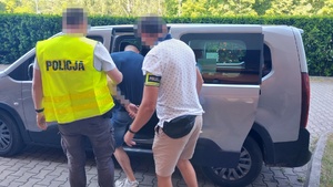 Policjanci asekurują zatrzymanego mężczyznę z kajdankami na nadgarstkach rąk trzymanych z tyłu, podczas wsiadania do samochodu.