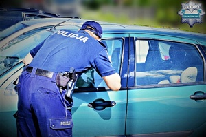 Policjant zagląda przez szybę do wnętrza pojazdu. Pojazd stoi w słońcu.