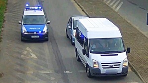 Po drodze jadą pojazdy. Po prawej biały bus za którym jedzie auto osobowe. po lewej policyjny radiowóz zapalonymi sygnałami uprzywilejowania.