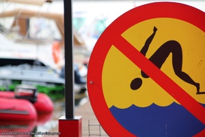 Znak zakazujący skoki do wody. W tle niewyraźne zarysy łodzi motorowych i żaglówek.