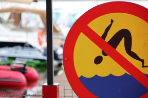 Znak zakazujący skoki do wody. W tle niewyraźne zarysy łodzi motorowych i żaglówek.