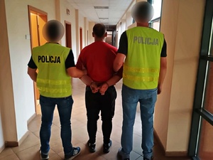 Policjanci w kamizelkach odblaskowych, prowadzą korytarzem zatrzymanego mężczyznę.