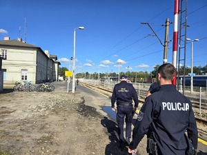 Umundurowani funkcjonariusze Policji i Strażnik Ochrony Kolei idący wzdłuż torów kolejowych na dworcu.
