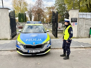 Radiowóz policyjny oznakowany i funkcjonariuszka Policji na tele cmentarza.
