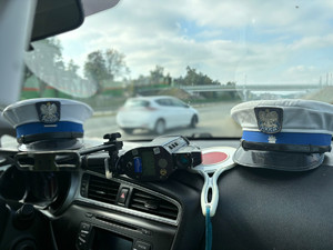 Wnętrze radiowozu policyjnego. Na desce rozdzielczej leżą czapki policyjne, urządzenie służące do pomiaru prędkości oraz sygnalizator służący do zatrzymywania pojazdów.