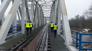 Umundurowani funkcjonariusze Policji i Służby Ochrony Kolei patrolujący tory na moście.